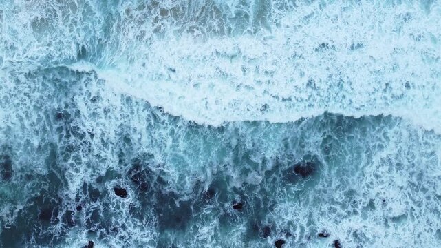 Aerial ocean footage of a broad blue wave rolling toward the shore, with textured foam, deep water tones, and powerful natural motion.