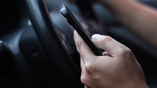 Close up of hands holding a smart phone at a car steering wheel, illustrating distracted driving, mobile phone use and road safety risks.