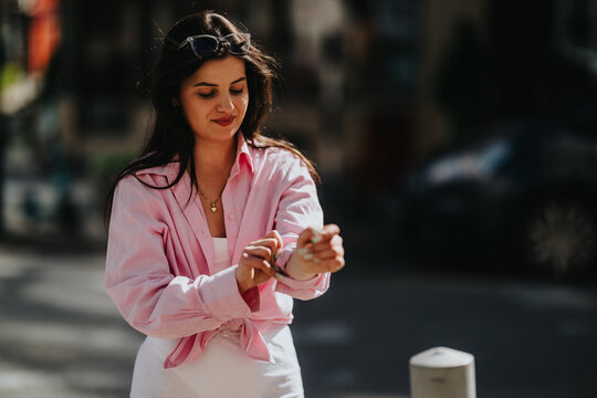 A young woman wearing a pink shirt and sunglasses adjusts her sleeve while smiling. She stands on a sunlit urban street in a casual, relaxed pose.