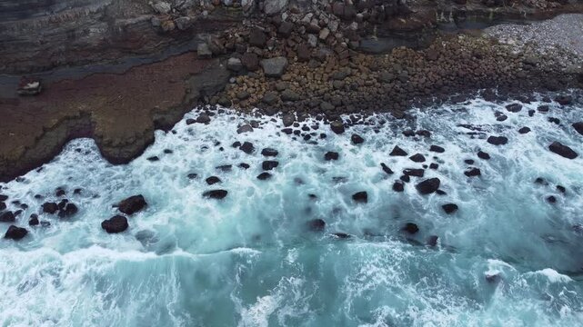 Drone footage of powerful turquoise waves breaking over a rocky shoreline, revealing dark stones, white foam, and dynamic coastal textures.