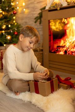 Cute preschool boy, sitting in front of the fireplace, holding christmas present and reading a book, joy and happiness