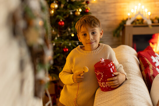 Cute preschool boy, sitting in front of the fireplace, holding christmas present and reading a book, joy and happiness