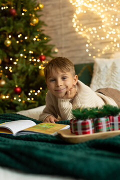 Cute preschool boy, sitting in front of the fireplace, holding christmas present and reading a book, joy and happiness