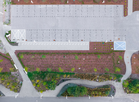 Aerial view of a modern paved parking lot with designated spaces, surrounded by landscaped garden beds with trees and shrubs, and pedestrian walkways in Sopot, Pomeranian Voivodeship, Poland.