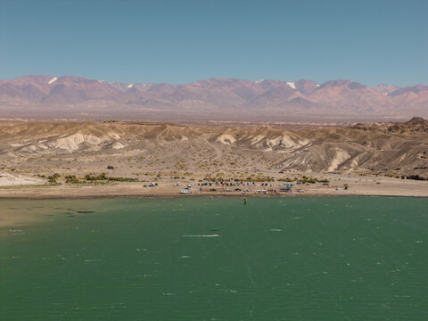 Aerial view of Cuesta del Viento reservoir with kiteboarders on the turquoise water, surrounded by arid mountains and desert landscape in Rodeo, San Juan, Argentina.