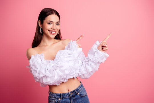 Smiling young woman in ruffled white blouse and jeans pointing to the side on a pink background, confident fashion portrait expressing happiness and playful style