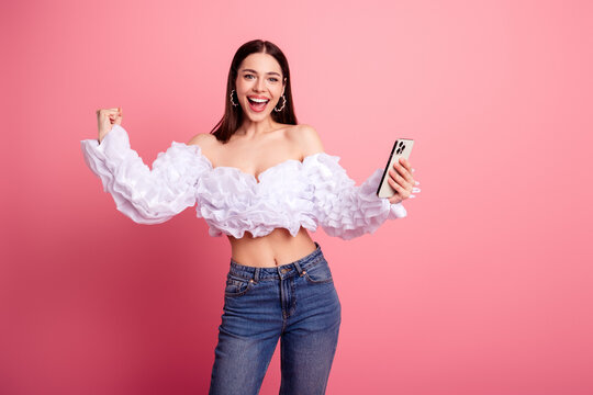 Excited young woman wearing a ruffled white off-shoulder top and jeans holding a smartphone and celebrating a joyful win with a big smile against a pink background