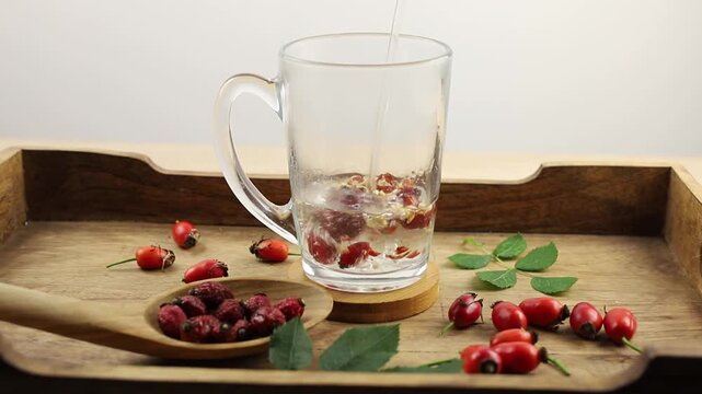 Making rose hip berry herbal tea, pouring boiling water over dried rose hip berries in a tea cup.Rose hip berries on wood spoon, decorated with fresh berries and green rose hip leaves, studio.