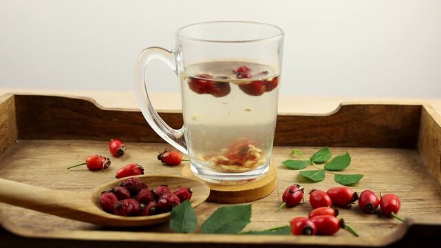 Making rose hip berry herbal tea, pouring boiling water over dried rose hip berries in a tea cup.Rose hip berries on wood spoon, decorated with fresh berries and green rose hip leaves, studio.