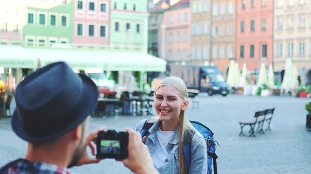 A man wearing a hat photographs a smiling blonde woman with a blue backpack in a colorful urban market square filled with outdoor furniture and potted plants.