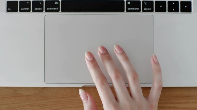 Top view close up shot of female hand with long natural manicured nails tapping and scrolling on laptop aluminum trackpad touch pad on wooden desk surface