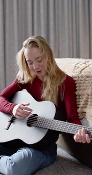 Vertical video: Woman holding white guitar looking down, plucking and singing, practicing at home