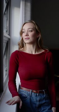 Vertical video: Adult female standing by home window, soft light drawing gaze, hand on sill