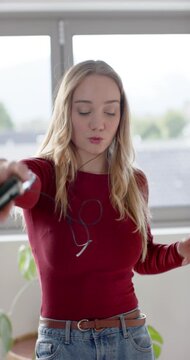 Vertical video: Woman at window with wired earphones and jeans, holding phone, dancing to music