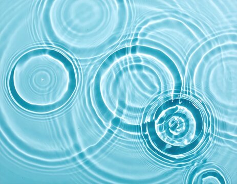 Concentric ripples radiating from multiple points on the surface of calm blue water, creating overlapping circular wave patterns and liquid textures.