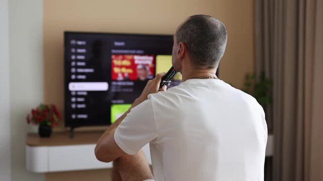 Young man sitting in front of TV and choosing what to watch using remote control