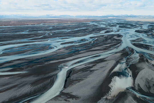 Aerial view of a braided glacial river system with light blue water flowing through dark volcanic sand plains in Skaftarhreppur, Iceland.