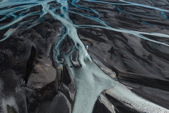 Aerial view of a kite surfer gliding over the turquoise glacial waters of a braided river winding through the dark volcanic sand landscape of Skaftarhreppur, Iceland.