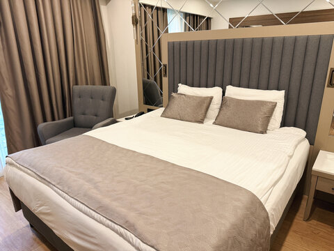Modern hotel room with double bed, white linen, beige pillows, bed runner, gray upholstered headboard, armchair, mirror panels, brown curtains, concept of hospitality, tourism, accommodation