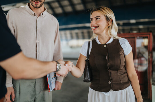 A smiling woman shakes hands with a man while another colleague stands nearby. The friendly introduction takes place on a covered transport platform, showing a casual business meeting.