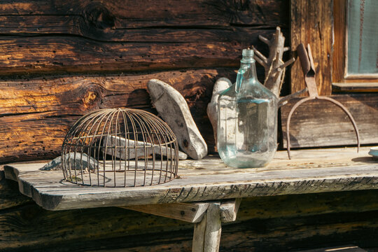 A collection of rustic vintage items including a metal cage, wooden shoemaker lasts, a large glass bottle, and an old oven peel rest on a wooden bench against a log wall.