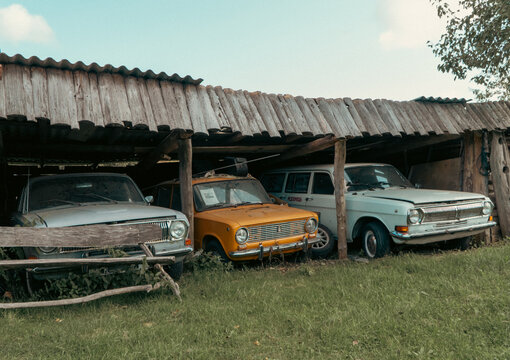 Minsk, Belarus - 08.13.2026: Three classic vintage Soviet automobiles sit parked under a weathered wooden barn shed on green grass in a countryside setting on a clear day.