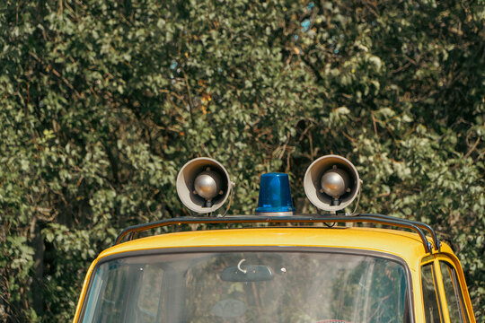 A vintage yellow service vehicle features a blue emergency flashing beacon flanked by two round loudspeaker horns mounted on its roof against green foliage.