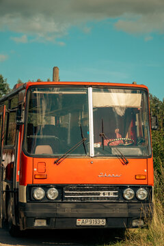 Minsk, Belarus - 08.13.2026: An iconic red vintage Ikarus coach bus manufactured in Hungary with dual round headlights and wipers stands parked outdoors against a clear blue sky and green trees.