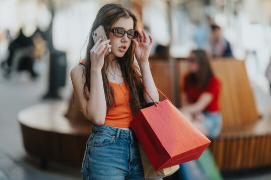 Young woman holds shopping bags and talks on her smartphone while adjusting sunglasses. Casual street shopping scene with blurred passersby in the background.