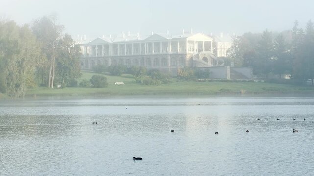 The Cameron Gallery stands gracefully through soft mist beyond a peaceful lake. Ducks swim gently on calm waters. This dreamlike view evokes quiet wonder in Catherine Park, Pushkin, Saint Petersburg.