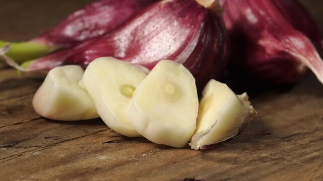 Macro shot of sliced garlic and whole purple cloves slowly moving on wooden surface. Close up of fresh raw organic garlic bulbs in studio light.