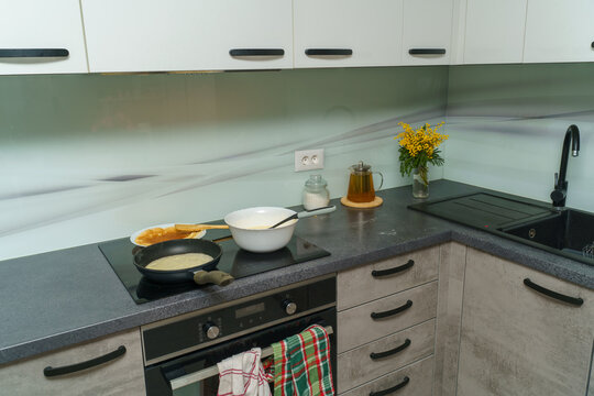 Spacious modern kitchen with pancakes being prepared on the cooktop, mixing bowl and wooden spatula on the counter, mimosa bouquet near the sink