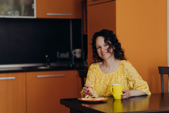Smiling woman in yellow dress sits with bright mug at kitchen table creating calm coffee break scene connected with self care mindful pause emotional wellbeing slow mornings and work life balance.