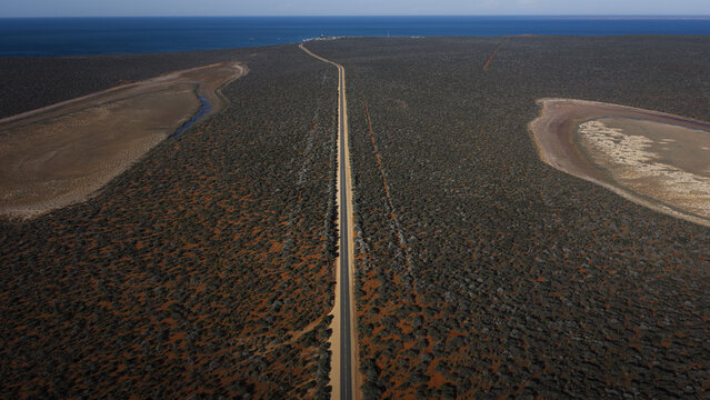 Aerial view of a straight asphalt road cutting through the arid, shrub-covered landscape of Western Australia leading towards the blue ocean in Perth, Western Australia, Australia.