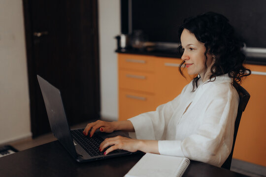 Smiling woman works on laptop at kitchen table creating home office scene linked with freelance careers digital inclusion flexible work schedules online business confidence and modern work culture.