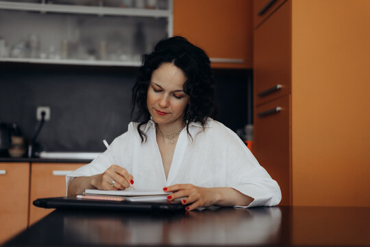 Woman writes notes beside laptop at kitchen table creating focused learning and planning scene linked with reskilling online education knowledge economy development productivity and lifelong learning.