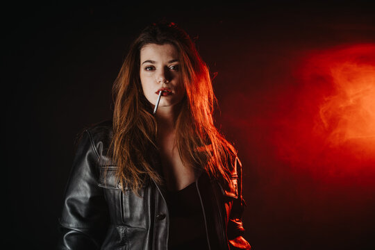 Young woman wearing a leather jacket looks directly at camera with a cigarette in her mouth. The moody portrait features red backlight and haze for an edgy studio look.