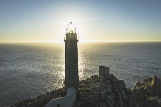 Aerial view of a stone lighthouse perched on a rocky cliff overlooking the Atlantic Ocean at sunset with golden light reflecting on the water in Galicia, Spain.