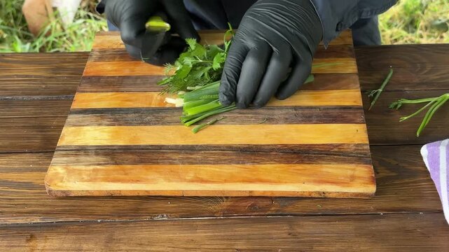 Close-up of a person wearing black gloves chopping herbs on a wooden board outdoors. High quality 4k footage