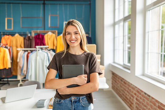 A young woman stands in her small clothing store, smiling while holding a tablet. The shop features colorful garments hanging in the background, with a bright and welcoming atmosphere.