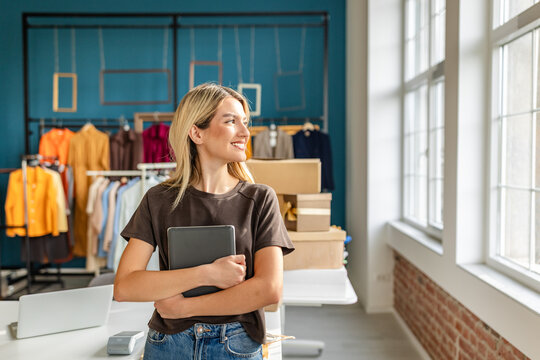 A young woman stands in her clothing business showroom, smiling while holding a tablet. The space features colorful garments displayed on racks, with natural light streaming through large windows, cre