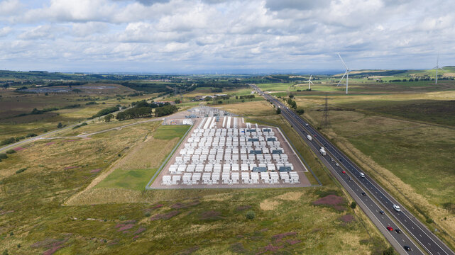 Aerial view of a large-scale battery energy storage system facility with rows of white containers situated in a rural landscape next to a highway in Coalburn, Scotland, United Kingdom.