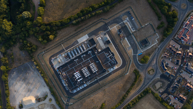 Aerial view of a large industrial facility with a fenced perimeter, parking lot, and surrounding residential area in Bracknell, England, United Kingdom.