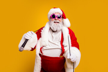 Obraz premium Santa Claus in star sunglasses holding a microphone and cane in a festive red fur-trimmed costume singing and partying against a bright yellow background