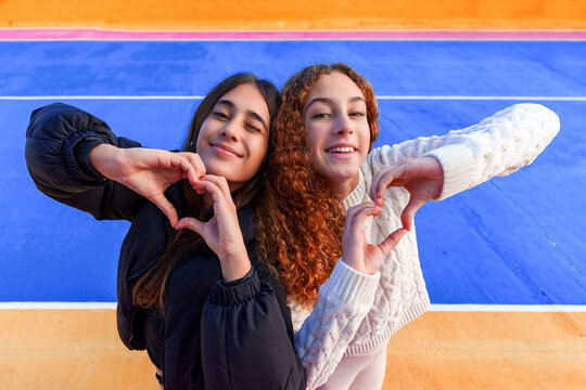 Two happy smiling young women making heart shapes with hands, showing friendship and connection on colorful background