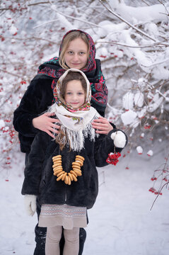 Two girls in traditional Russian folk headscarves playing in a snowy forest with ring-shaped bread and viburnum berries. The playful scene represents Russian folk culture and winter traditions.