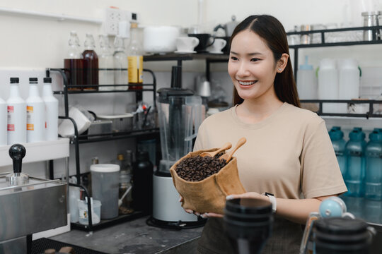 Smiling Asian barista holding burlap bag of roasted coffee beans in modern cafe, highlighting fresh ingredients, artisanal preparation, small business, hospitality and coffee shop.