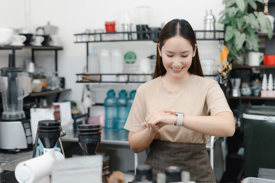 Smiling Asian barista checking time on smartwatch at coffee shop counter, surrounded cafe equipment, punctual service, time management, small business, workplace lifestyle.