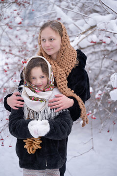 Two girls in traditional Russian folk headscarves playing in a snowy forest with ring-shaped bread and viburnum berries. The playful scene represents Russian folk culture and winter traditions.