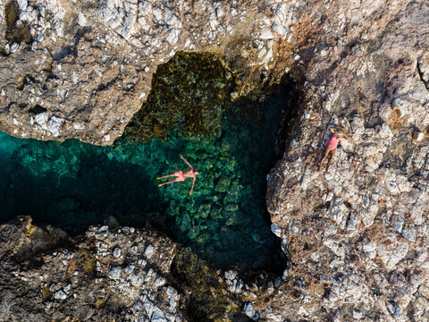 A person enjoys the clear turquoise waters while relaxing atop the rocky formations of Paros Island, Greece. Sunlight reflects off the water, creating a serene midday scene.